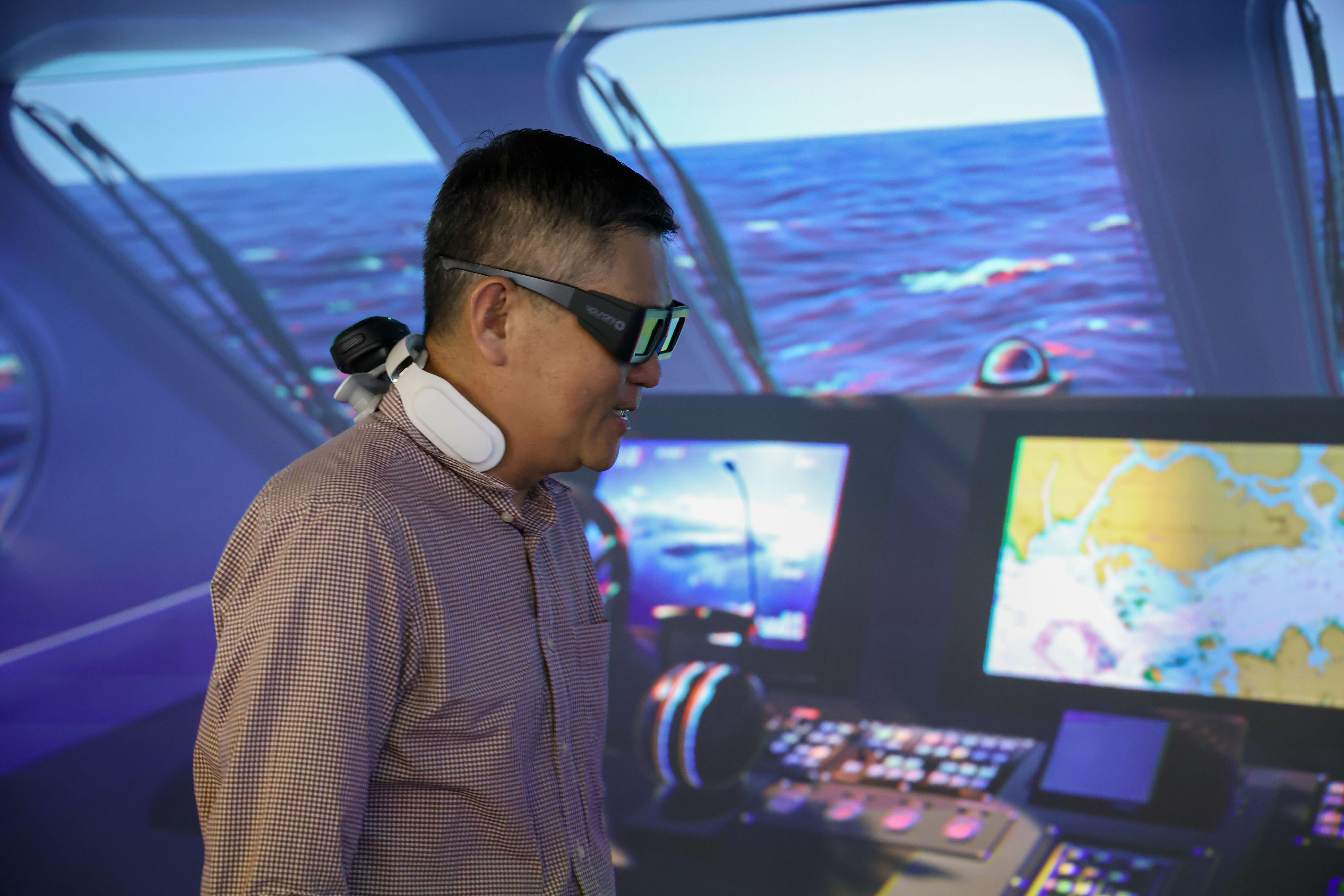 A man wearing VR goggles inside a simulated ship with multiple screens displaying navigational maps and ocean views.