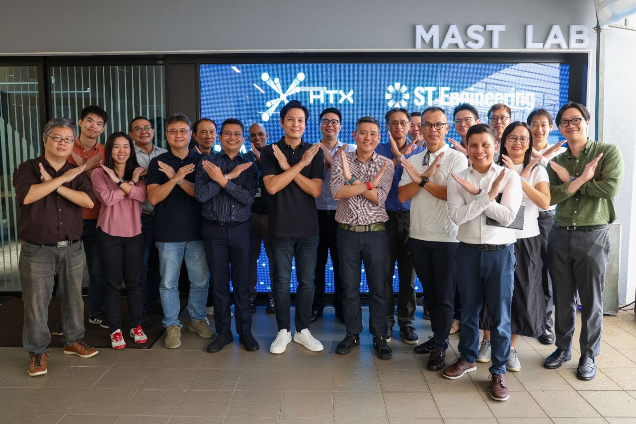 A group of 20 people standing in front of a digital display with HTX and ST Engineering logos, posing with arms crossed in an "X" shape.