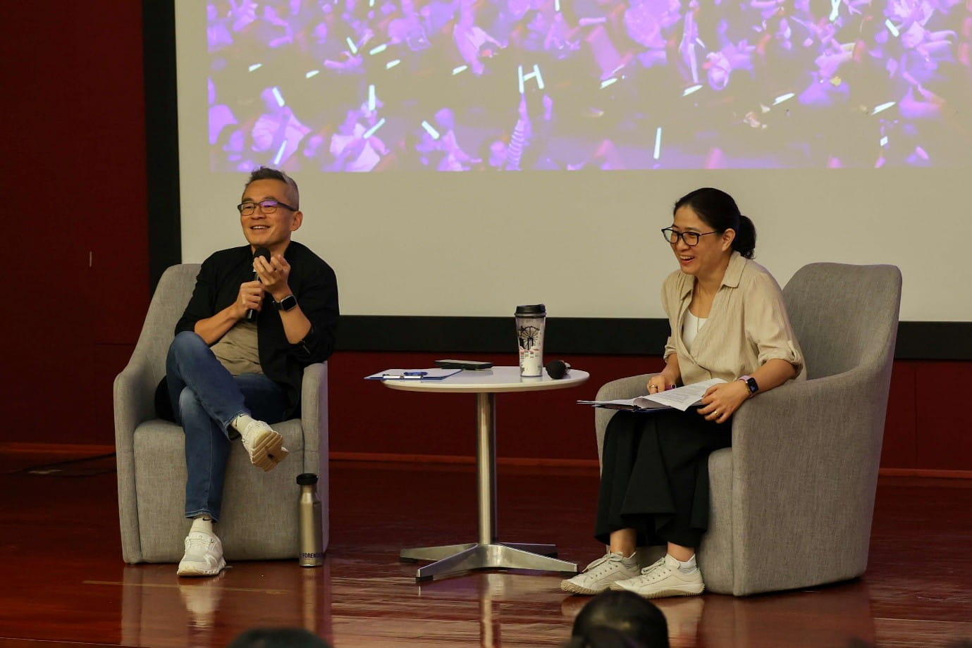 Two speakers seated in armchairs during a live discussion, with one holding a microphone and another holding notes.
