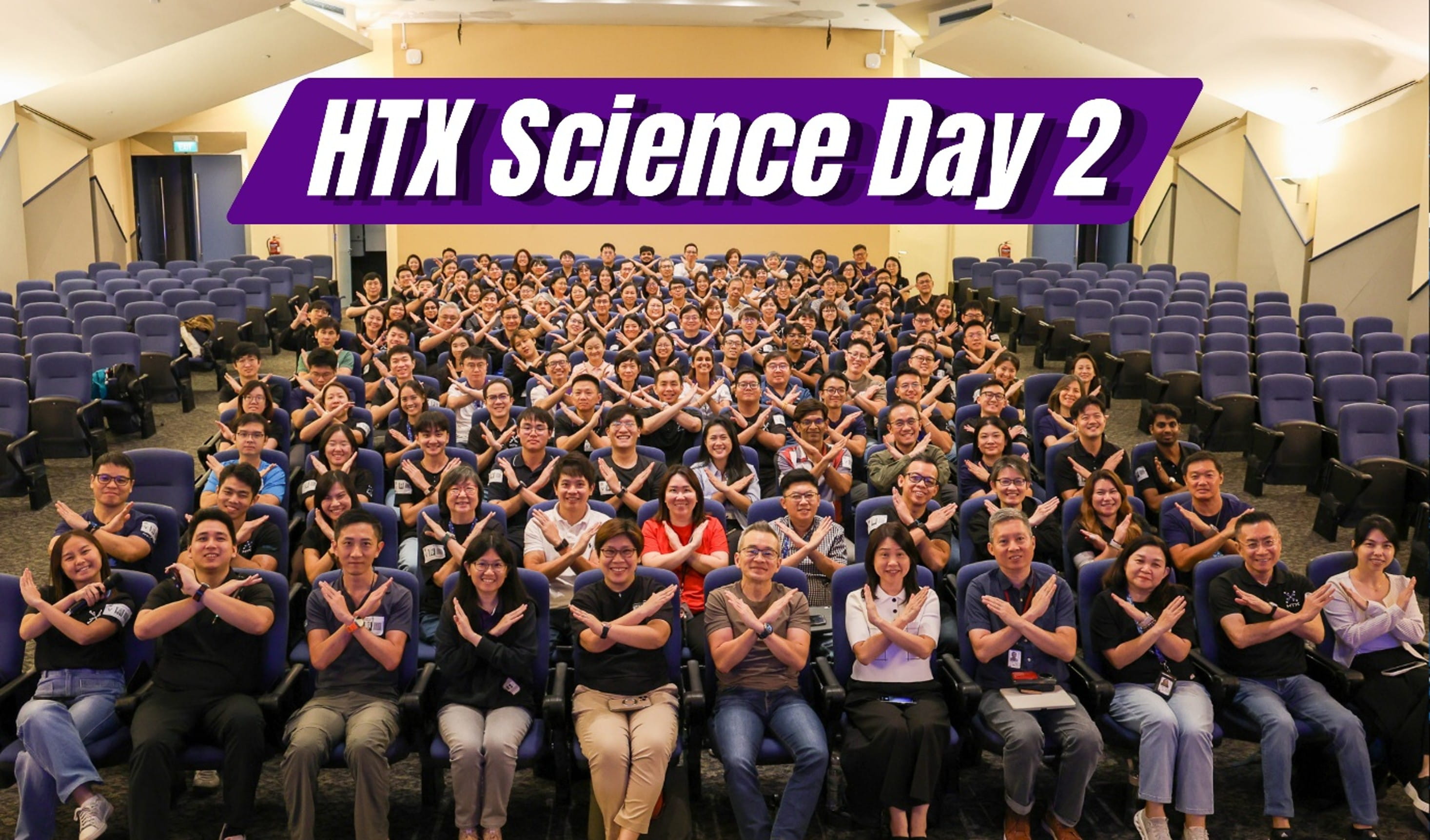 A large group of people seated in an auditorium, all posing with arms crossed in an “X” gesture. A purple banner with white text "HTX Science Day 2" is displayed above, indicating a science event.