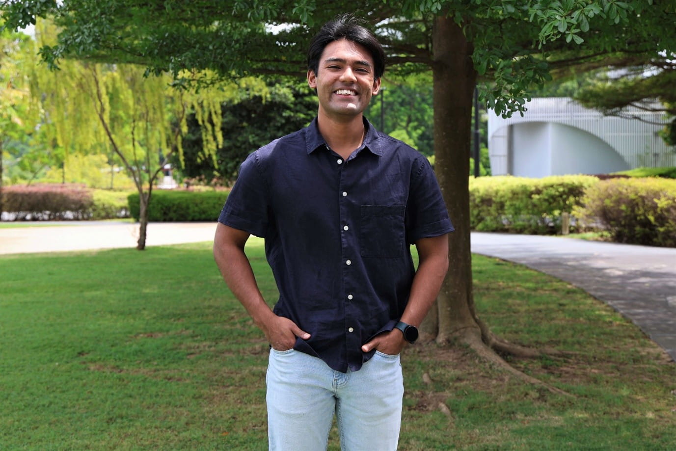 A man outdoors in a short-sleeve shirt, light jeans, and a wristwatch, with hands in his pockets.