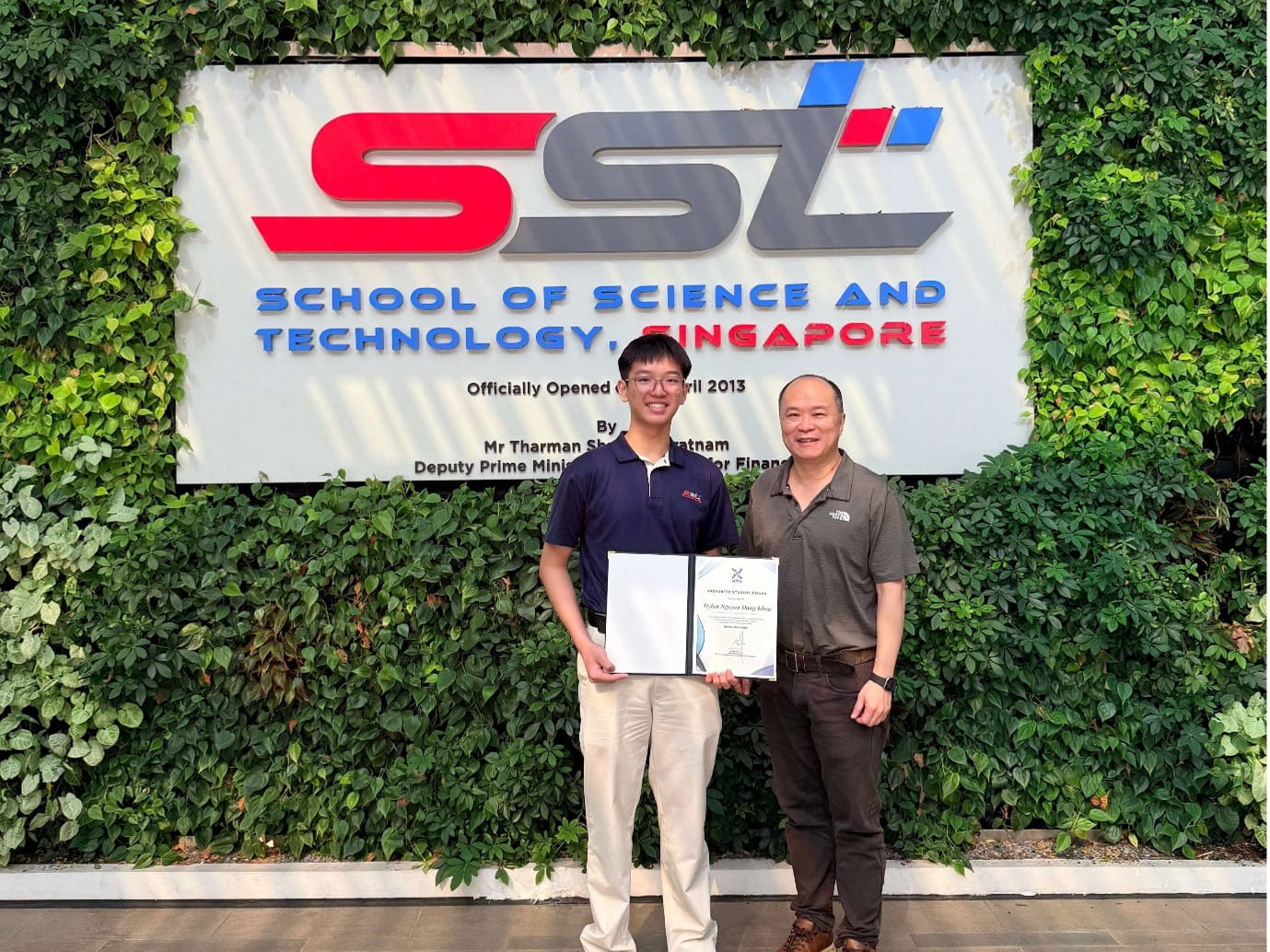 Two people pose in front of a School of Science and Technology sign. Younger man holds a certificate.