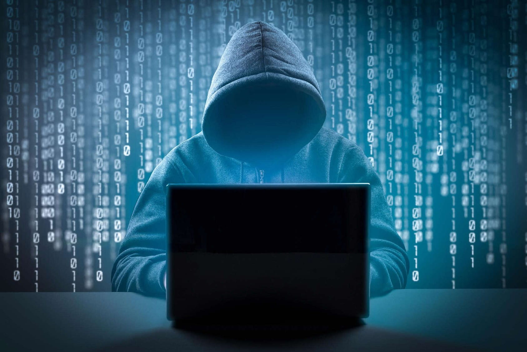 A person wearing a hoodie sitting behind a laptop with his face obscured, symbolising cybersecurity or hacking.