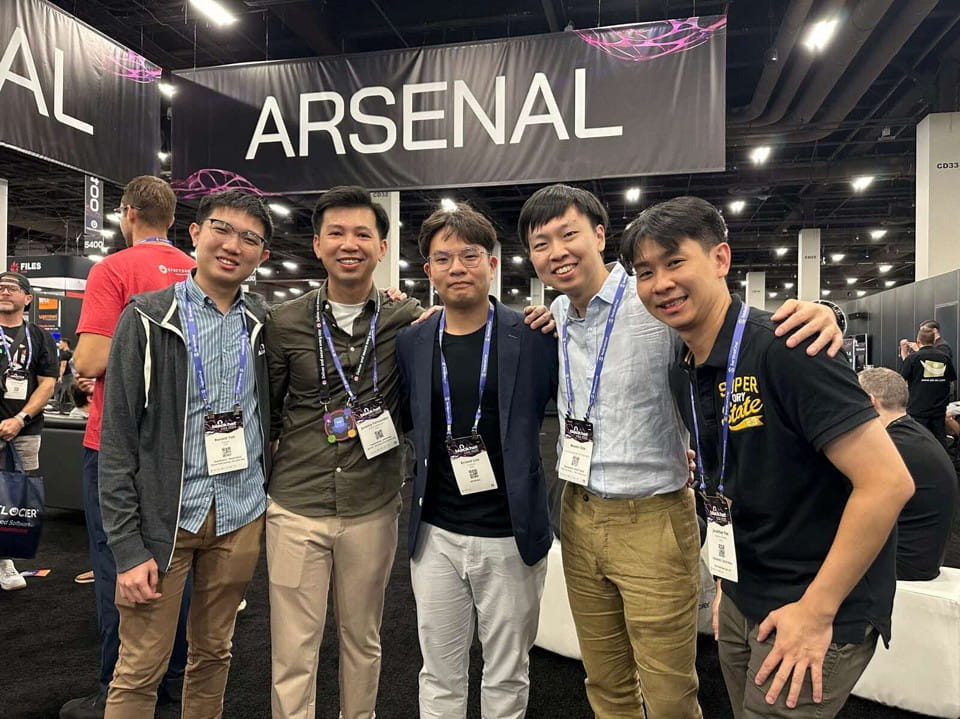 Five men standing together. All wear event lanyards. Background shows other attendees and booths.