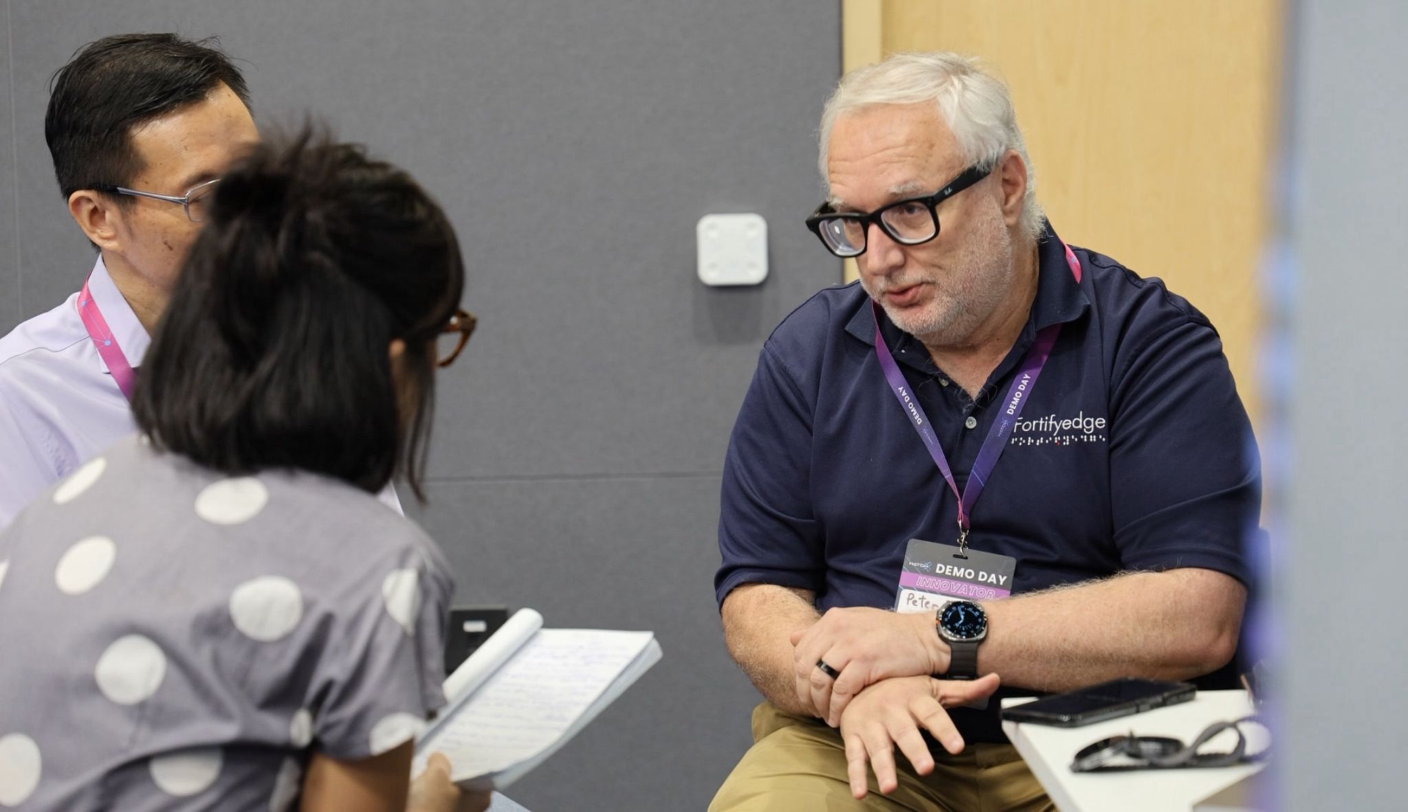 A man seated and speaking to a man and a woman holding a notebook.
