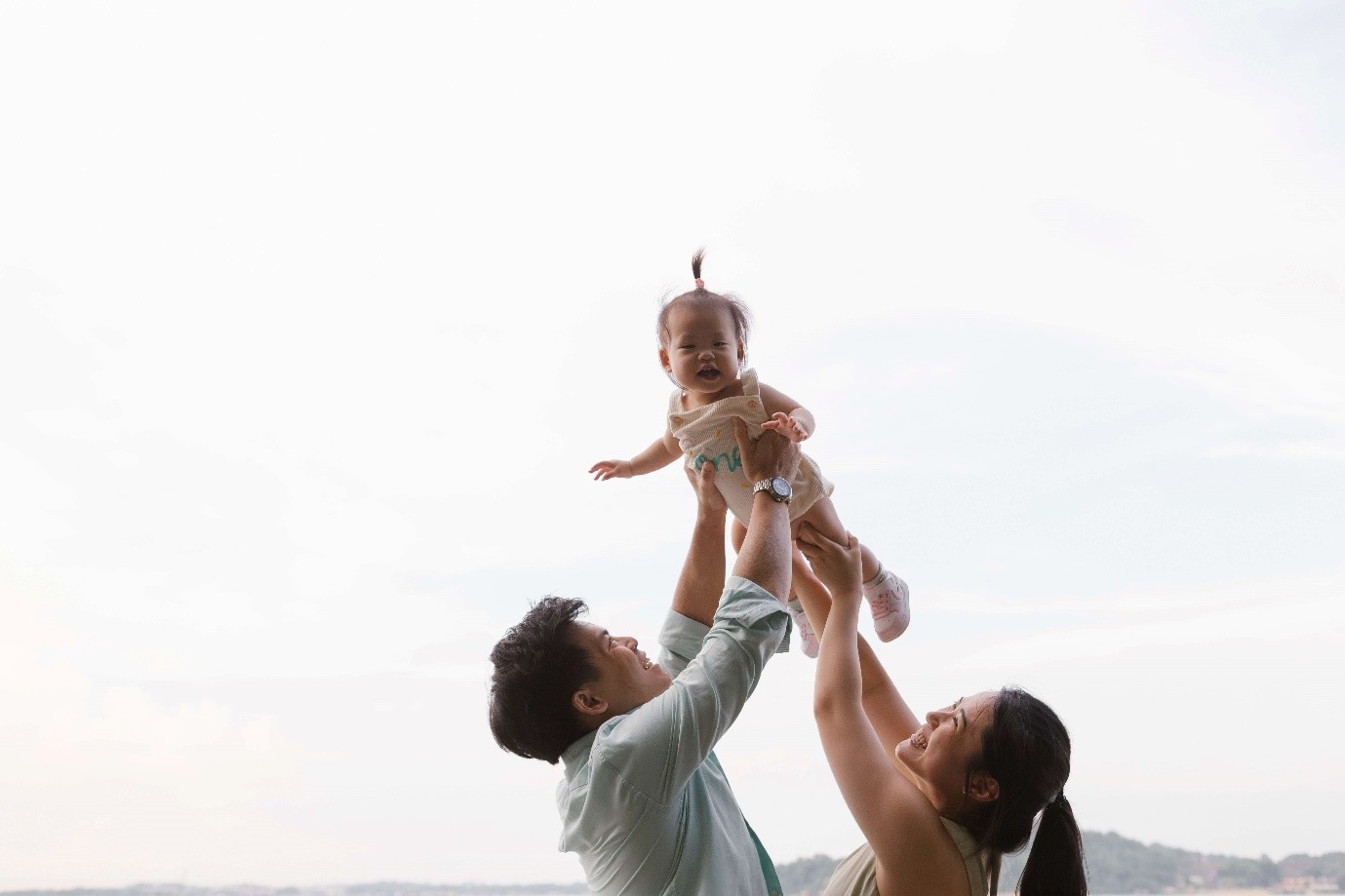 A happy couple lifts their baby into the air against the sky.