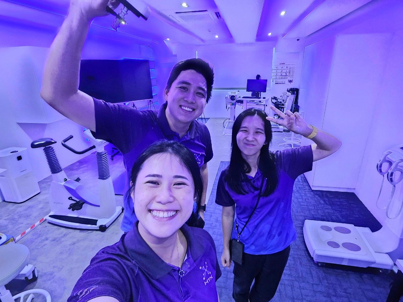 Three colleagues pose for a selfie in a room filled with scientific equipment and illuminated by purple lighting.