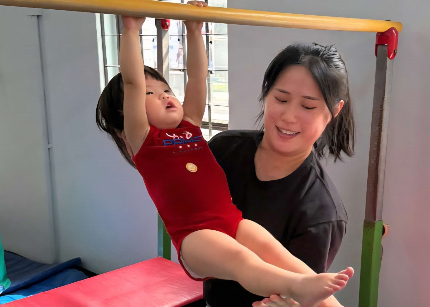 A woman supports a toddler as she practices hanging from a low wooden gymnastics bar.