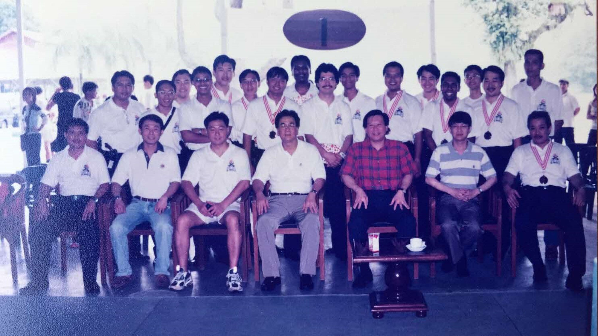 A large group of men in white polo shirts poses for a group photo, with a few men seated in the foreground.