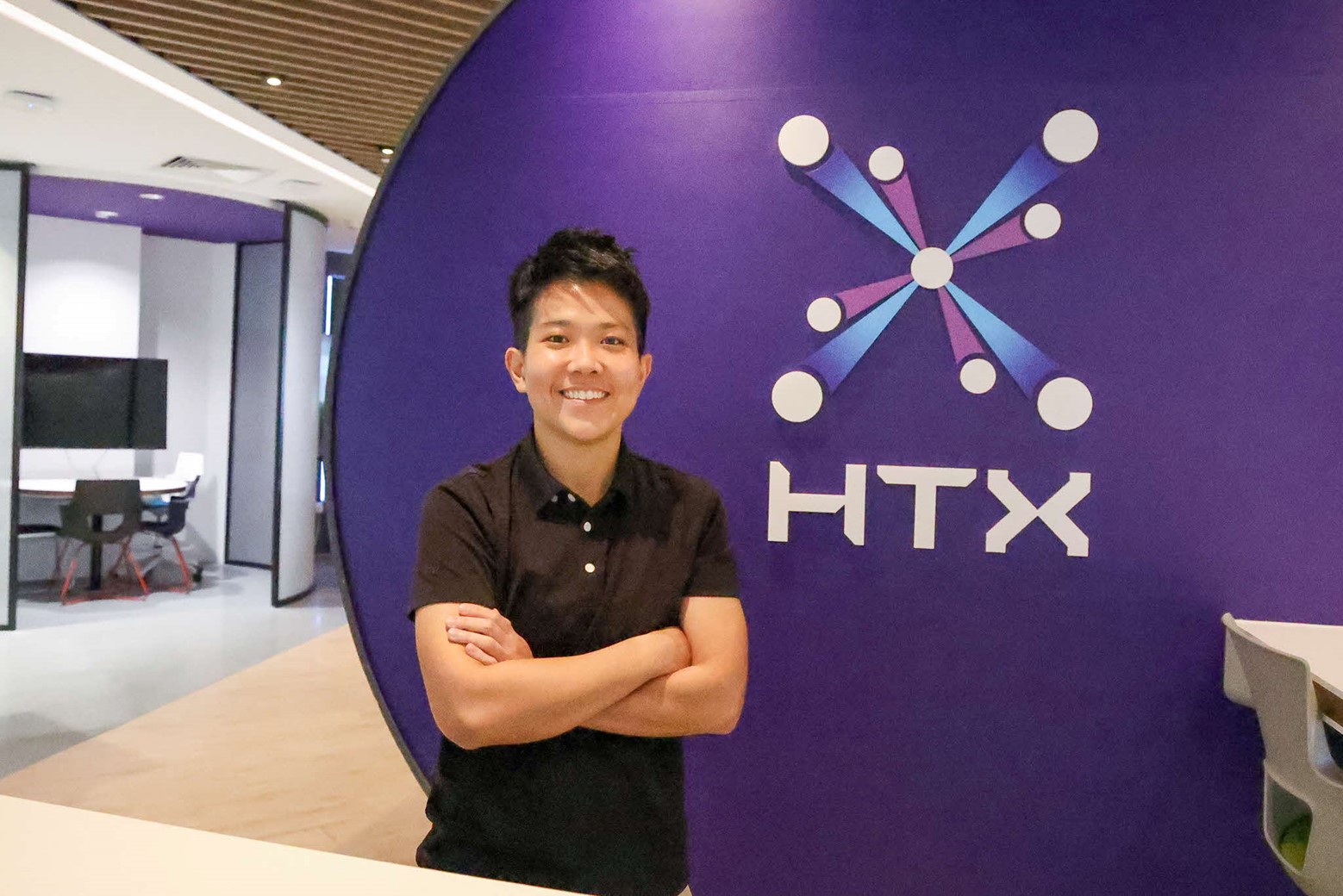 Priscilla Koh of HTX smiling with arms crossed in front of HTX logo in office