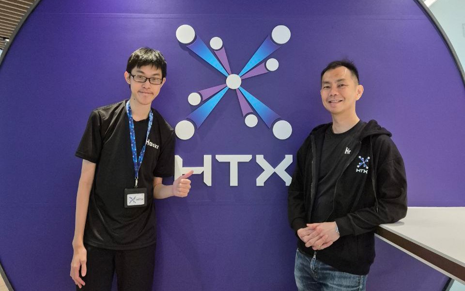 Two men posing for a photo in front of HTX’s logo.
