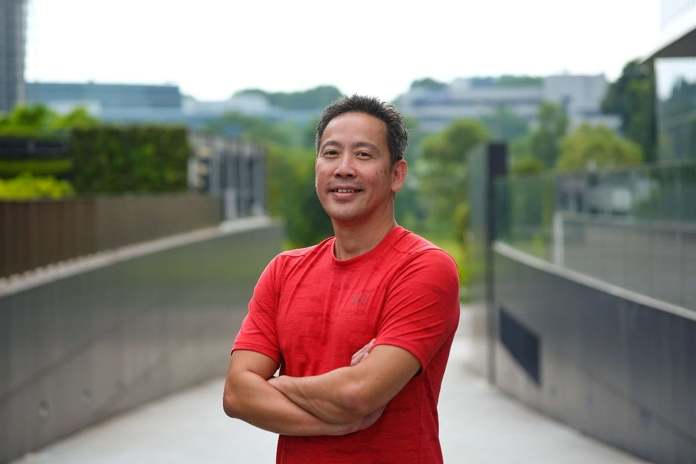 A man standing outdoors with arms crossed, wearing a red shirt and smiling at the camera.