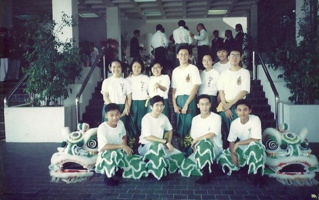 Two rows of young people posing. At the front row, one lion dance costume head sits at each side, while four youths don lion dance costume bottoms. At the back row, seven youths are dressed in matching white shirts and green pants.