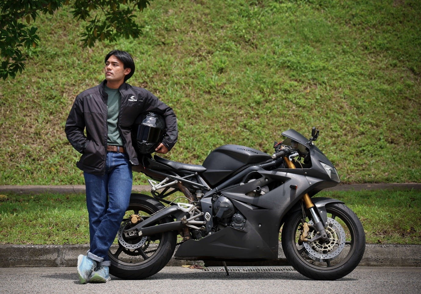 A young man in a riding jacket leans against a black sportbike on a roadside.