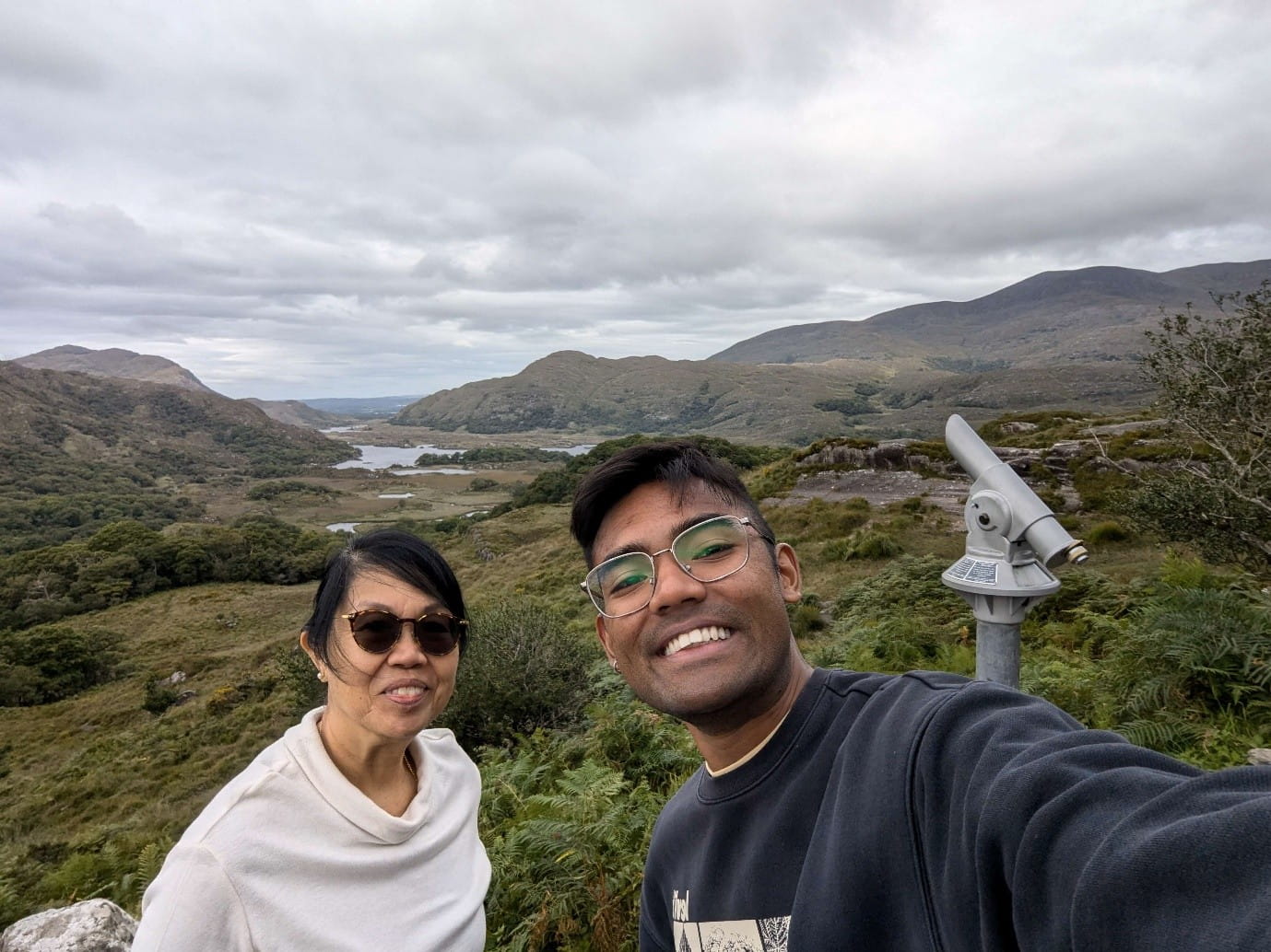 A selfie of a young man and a woman at a scenic mountain featuring lakes and a cloudy sky.