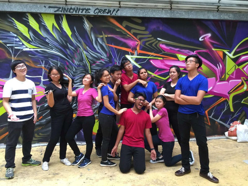A large group of people in colourful t-shirts strike playful and dramatic poses in front of a mural.
