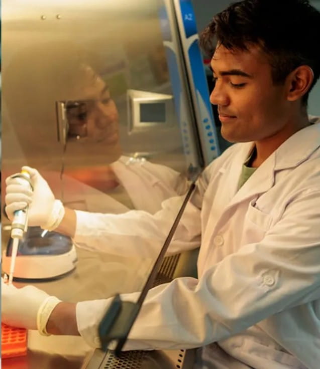A young man in a white lab coat and gloves uses a pipette inside a laboratory sterile workstation.