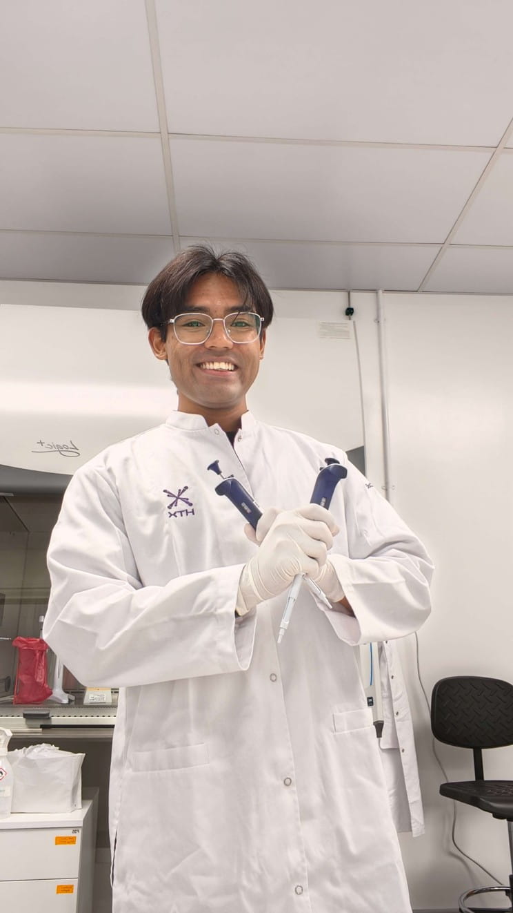 A smiling man in a lab coat poses while holding two laboratory pipettes crossed in front of him.