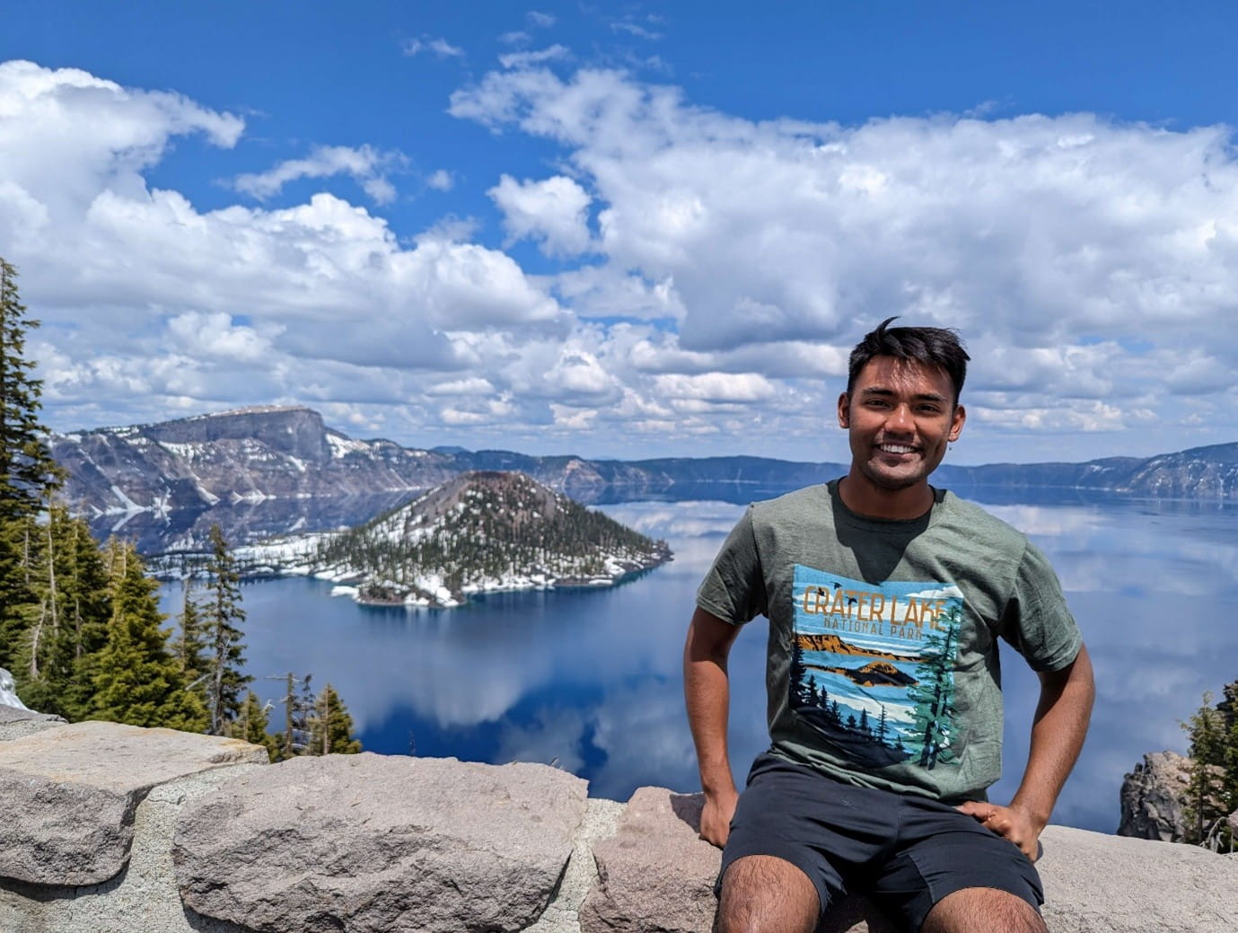 A young man sits on a stone wall overlooking the blue waters and volcanic island of Crater Lake.