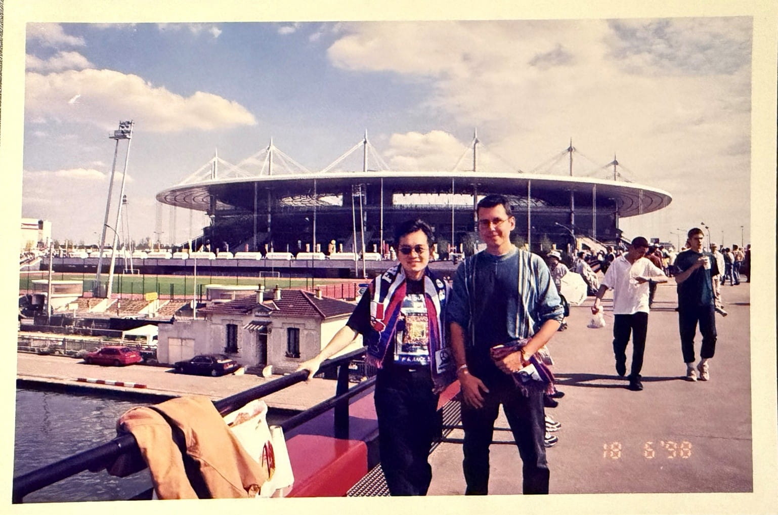 Two men standing along a walkway, against a backdrop of a large stadium with a circular roof.