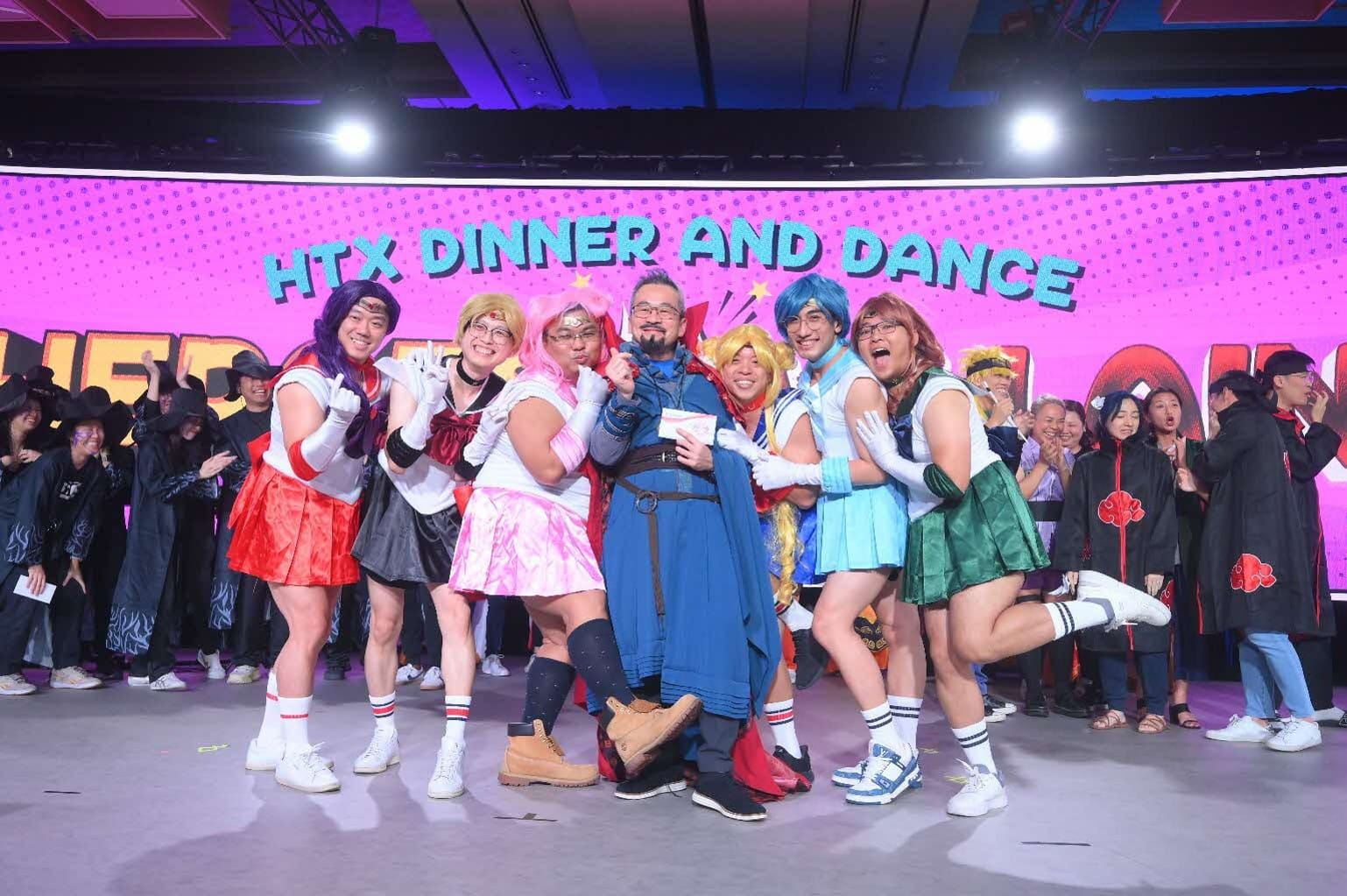 A group of seven people dressed in costumes posing against a backdrop reading "HTX Dinner and Dance".