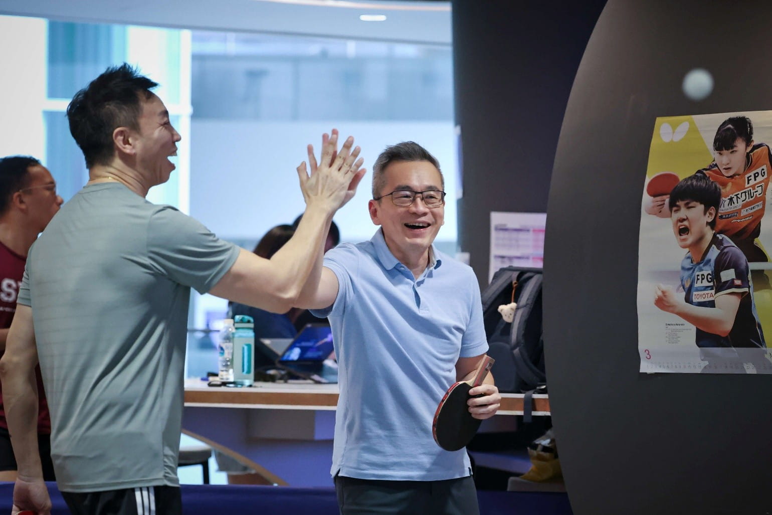 Two men celebrating with a high-five during an indoor table tennis match.