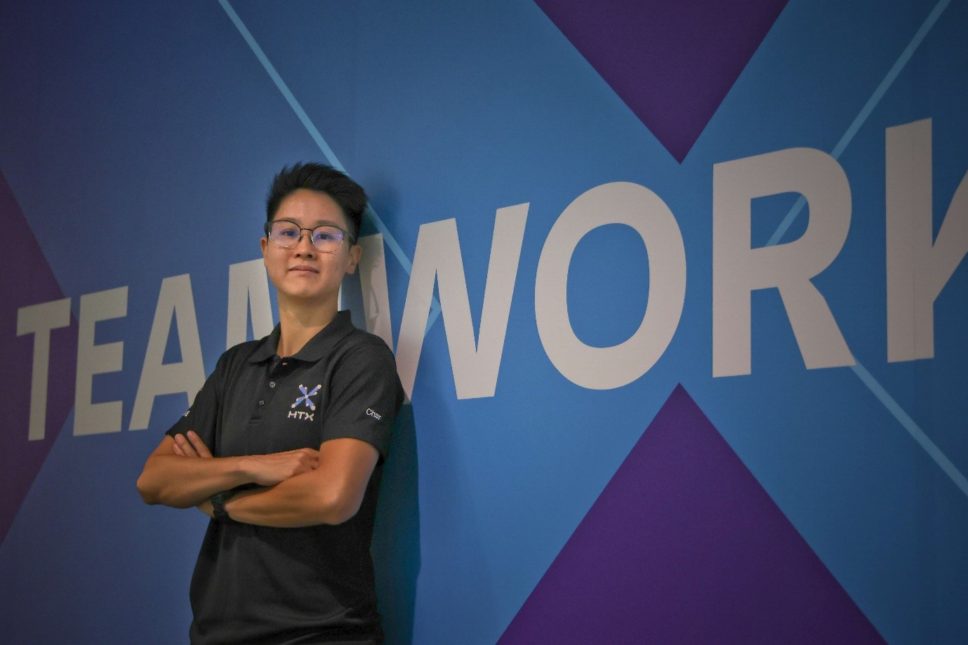 A lady in an HTX polo shirt leans against a wall.