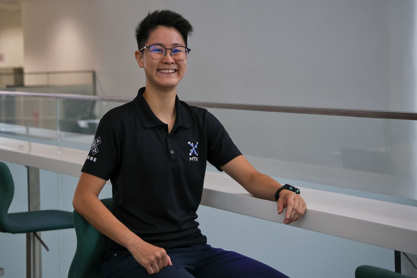 A smiling lady wearing a black HTX polo shirt sits at a white counter in a bright office environment.
