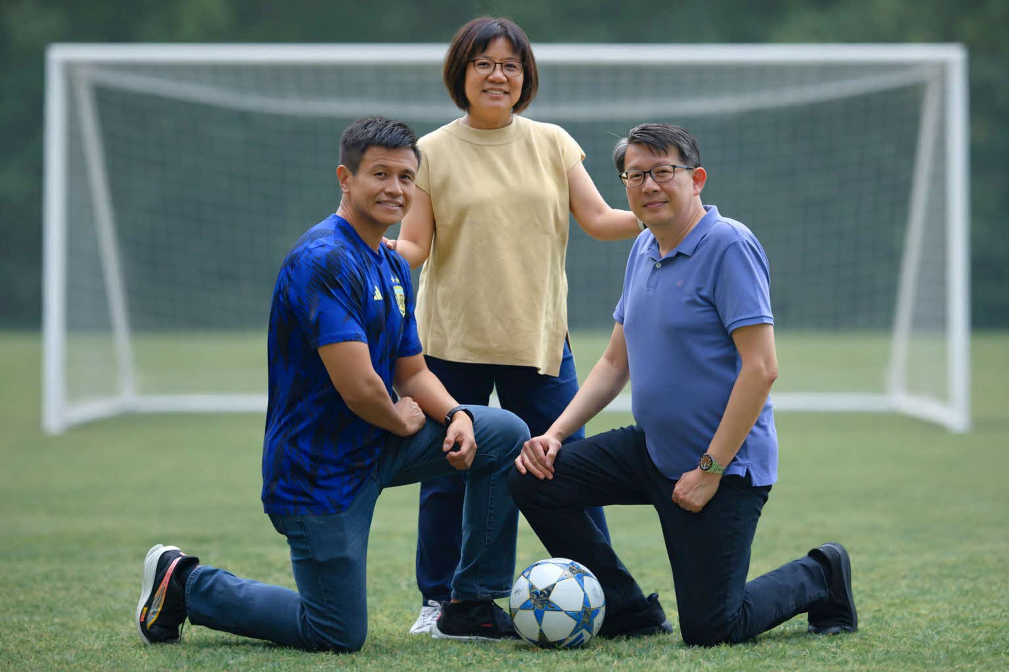Three individuals pose in front of a soccer goalpost on a grass field with a soccer ball at their feet.