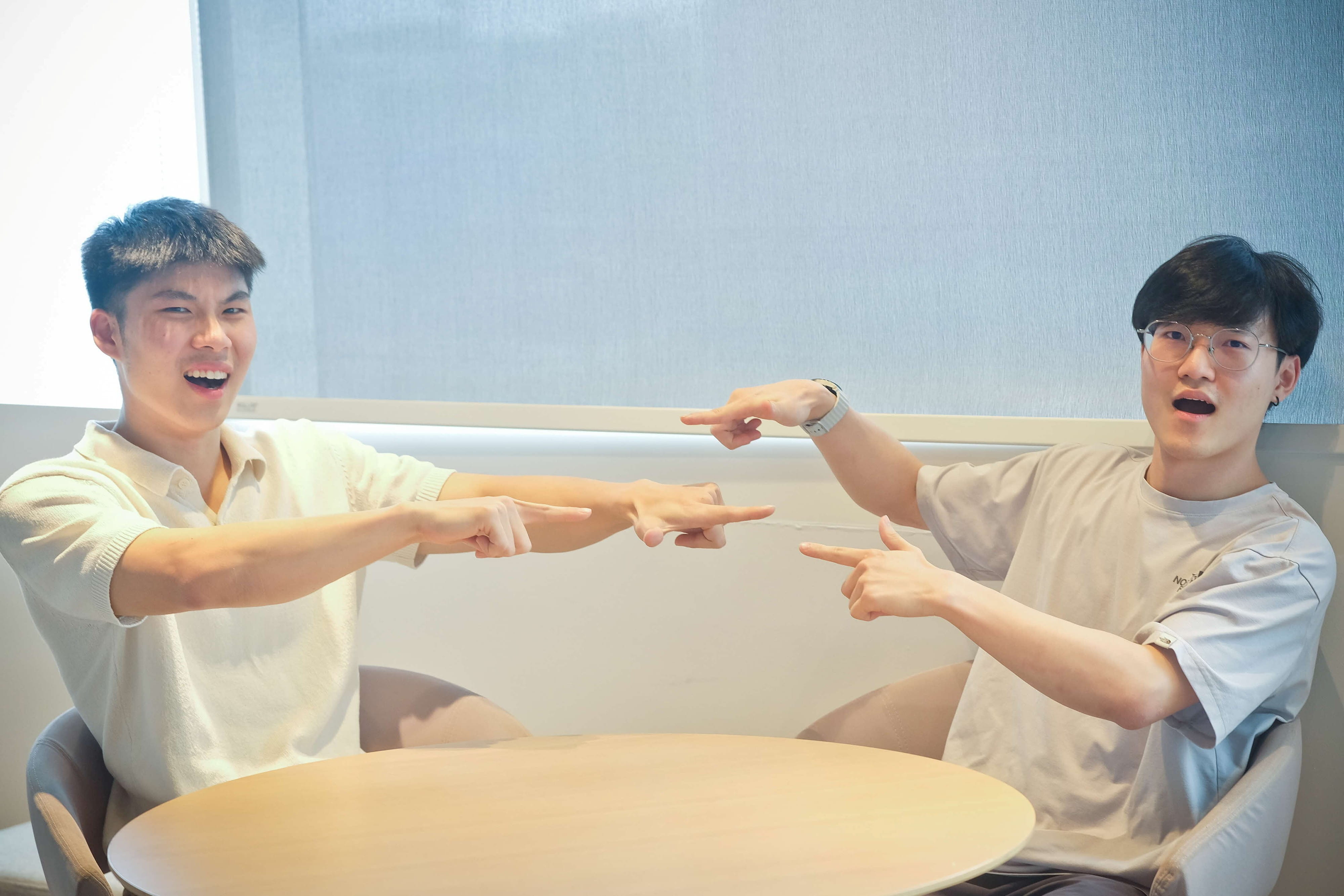 Two young men sitting at a round table in casual setting, both smiling and pointing at each other.