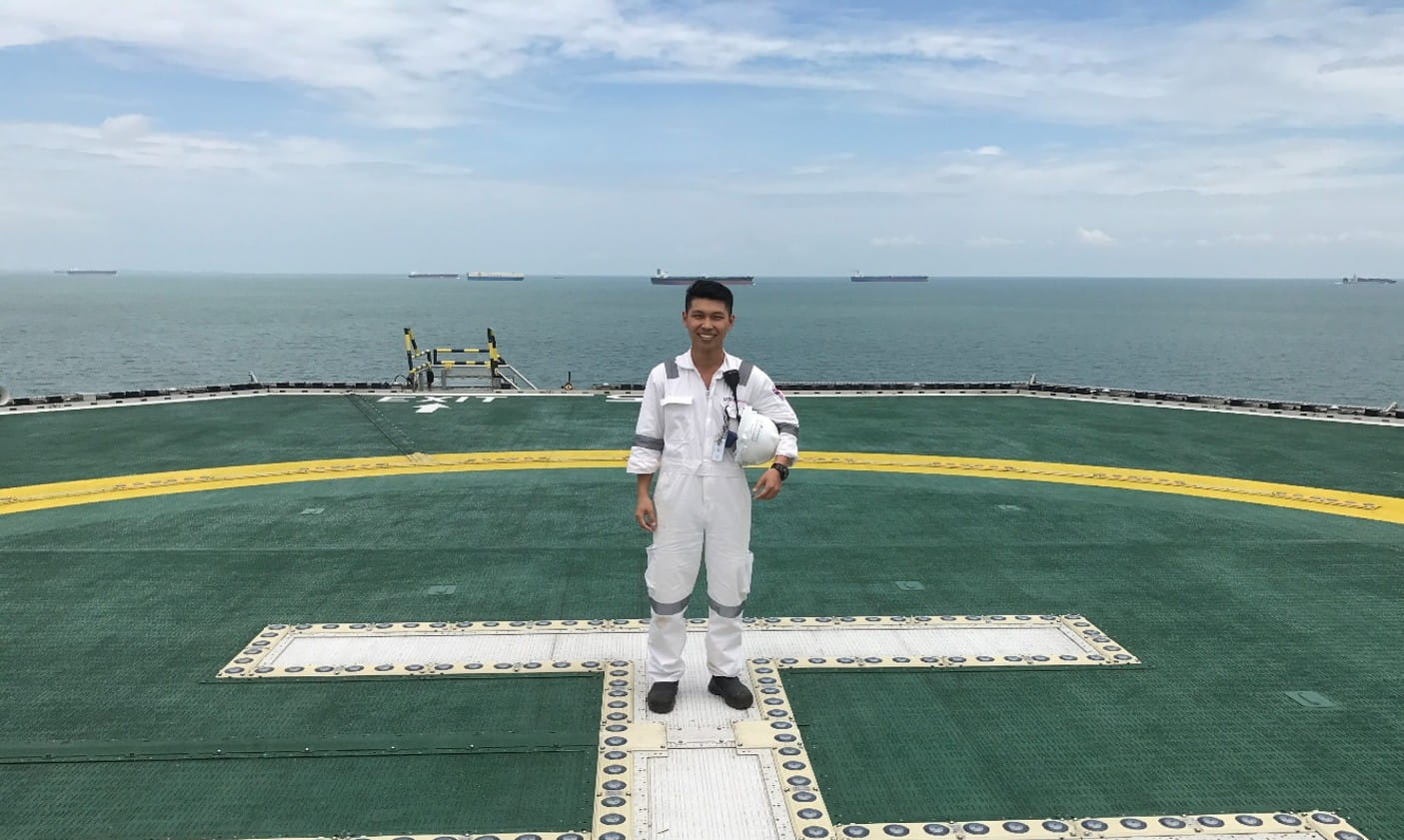 Man in white coveralls standing on a green helipad, with ocean and several ships visible in the background.