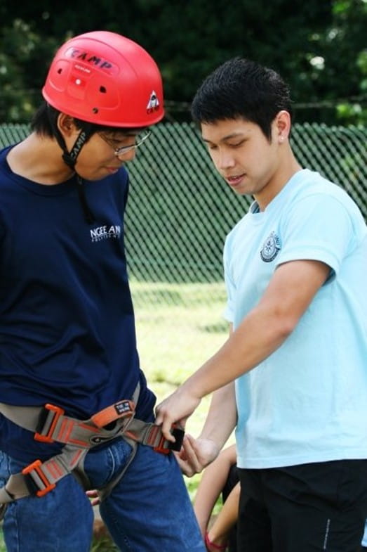 Two people outdoors, one wearing a red helmet and safety harness while Galvin assists with adjusting the harness.