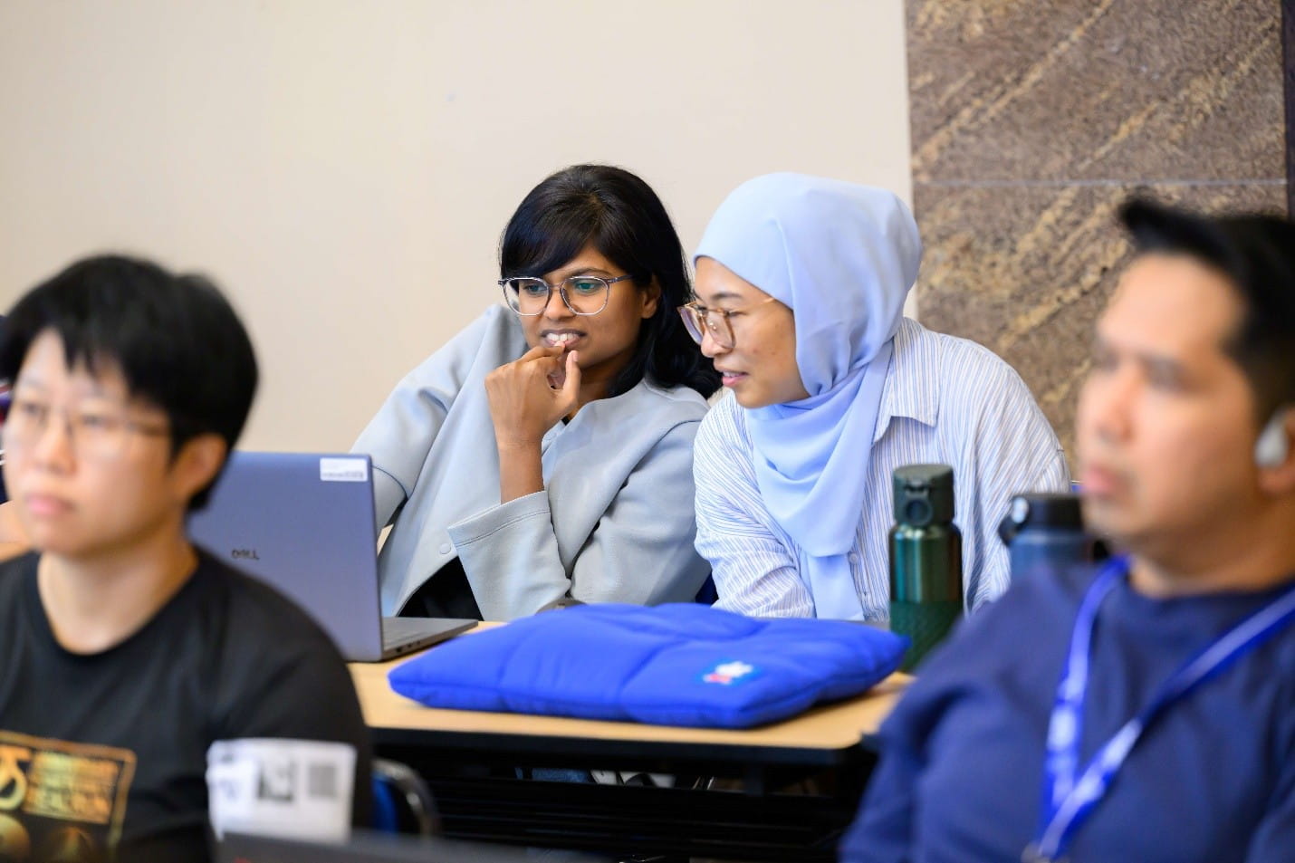 Two women sitting at a desk, looking intently at a laptop screen in a classroom setting.