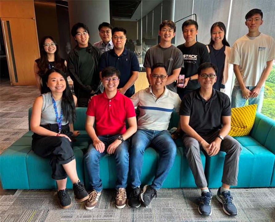 A group of 12 colleagues posing together on and around a teal sofa in a modern office environment.