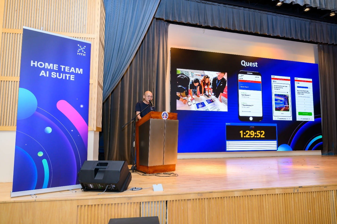 A man speaking at a lectern on stage next to a "Home Team AI Suite" banner, with a large screen displaying "Quest" features behind him.