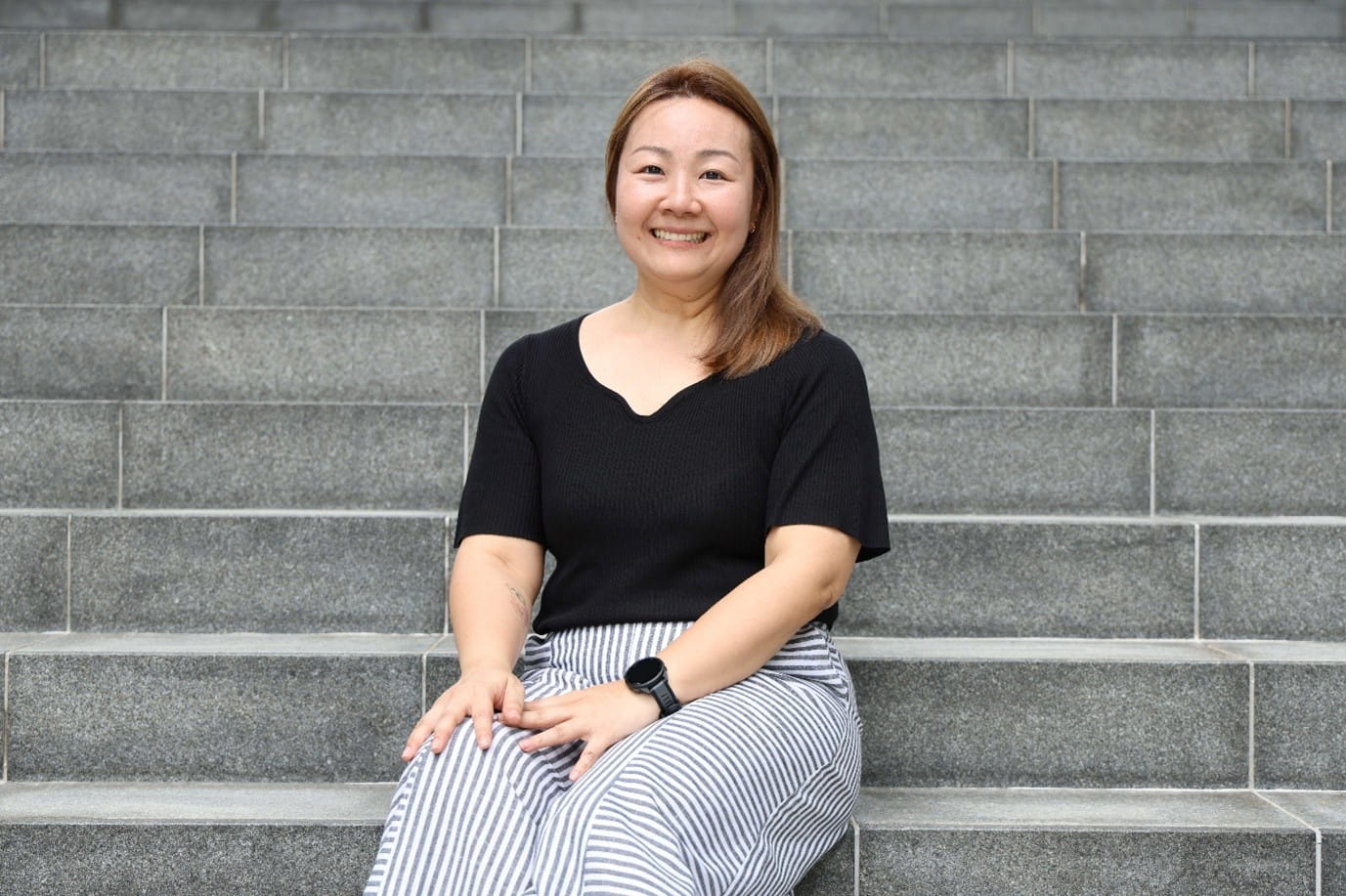 A woman sitting on outdoor stone steps wearing a black top and striped pants.