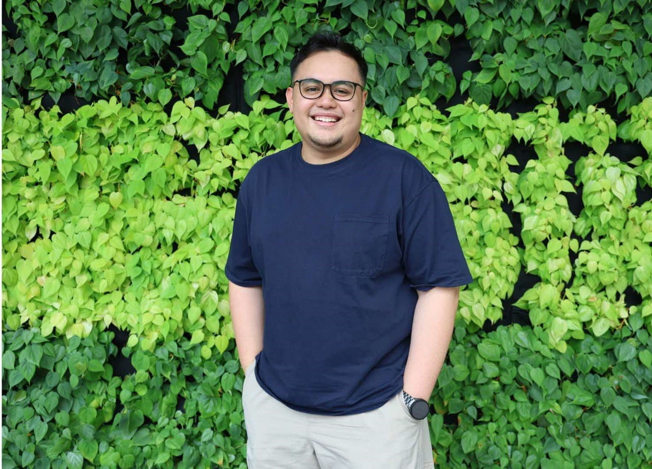 A man standing in front of a lush green leafy wall, wearing a blue t-shirt and light-coloured shorts.