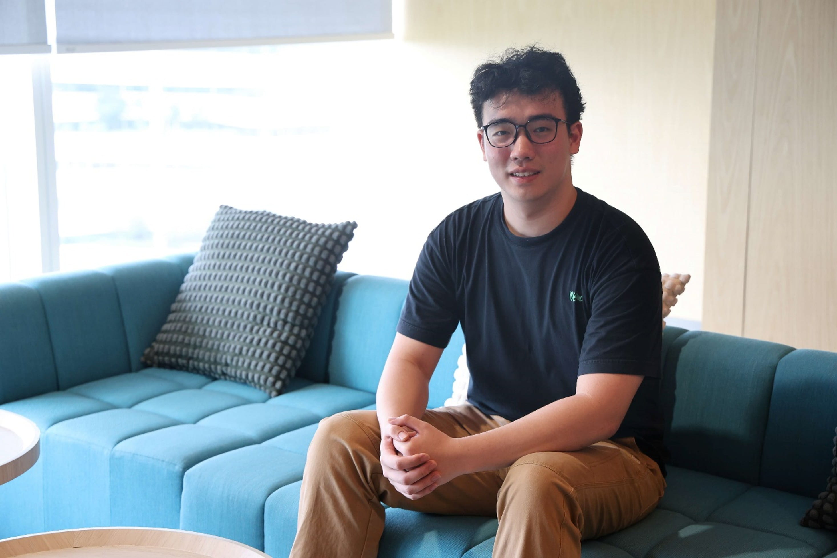 A young man wearing glasses, a black t-shirt, and brown pants sitting on a teal sofa.