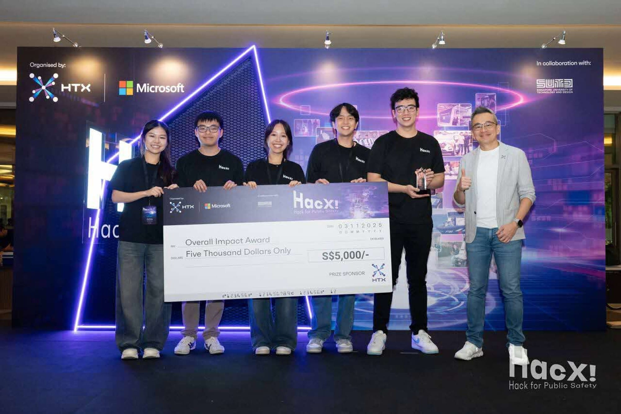 Five people on stage holding a large cheque for S$5,000 labelled "Overall Impact Award" from HacX!