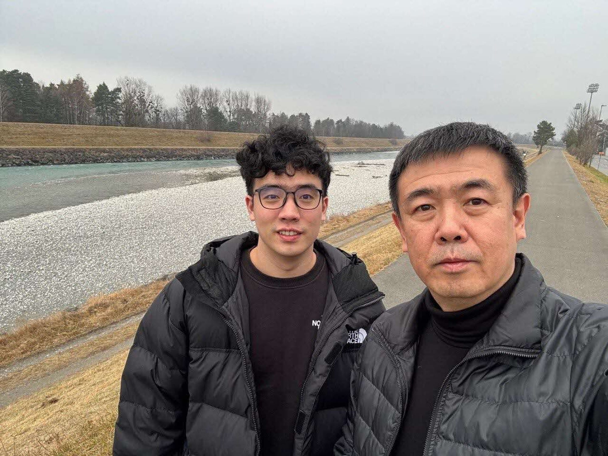 Selfie of a father and son in black jackets against an embankment and trees in the background.
