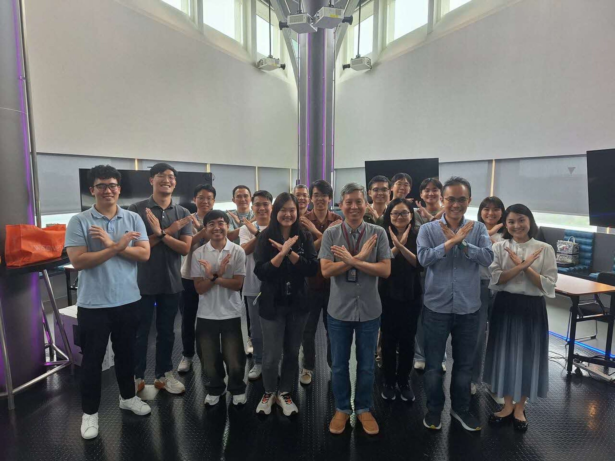 Large group of people standing indoors, with arms crossed in an "X" shape.