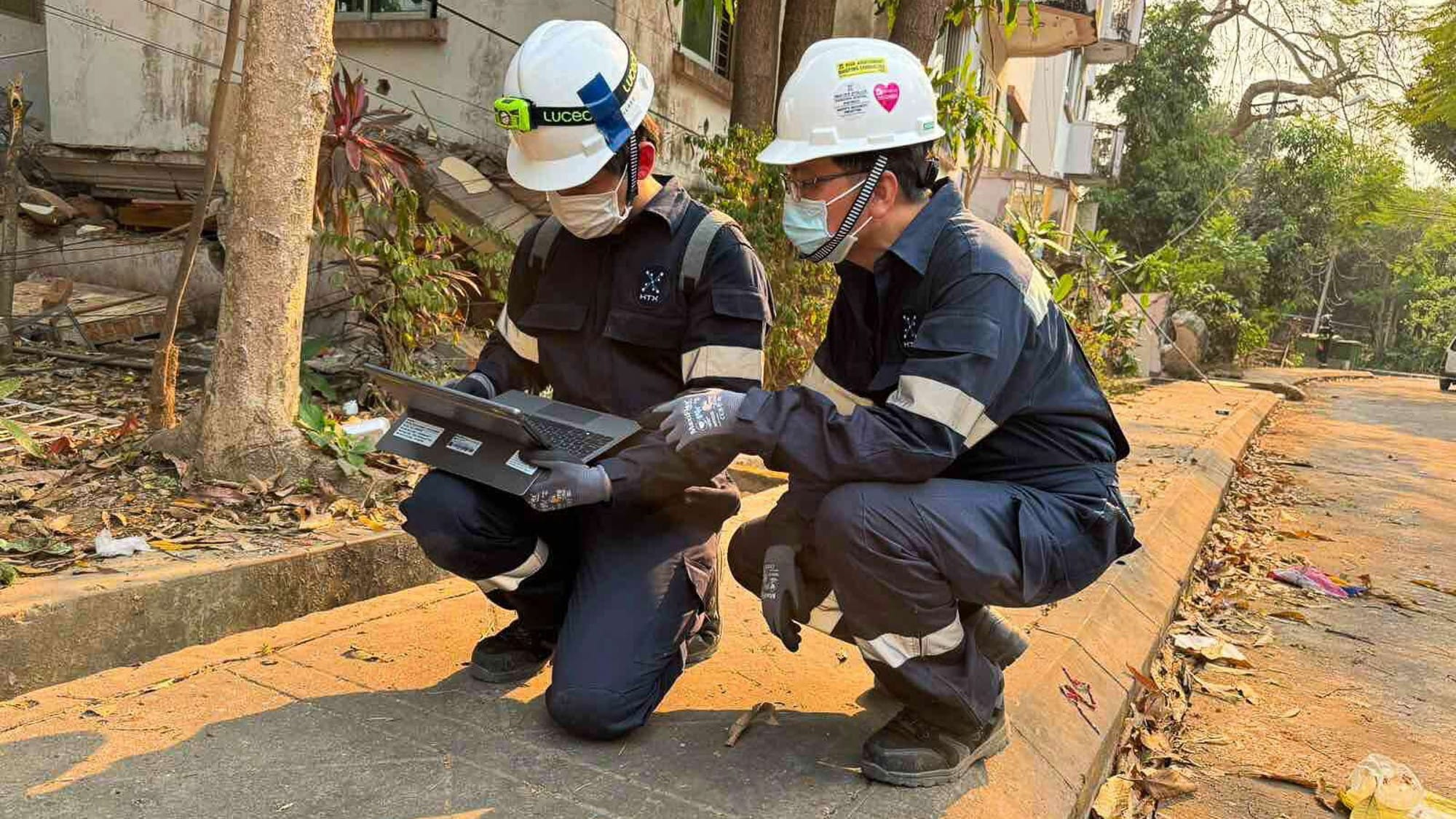 Two officers in helmets, masks, and protective uniforms examining equipment on a sidewalk.