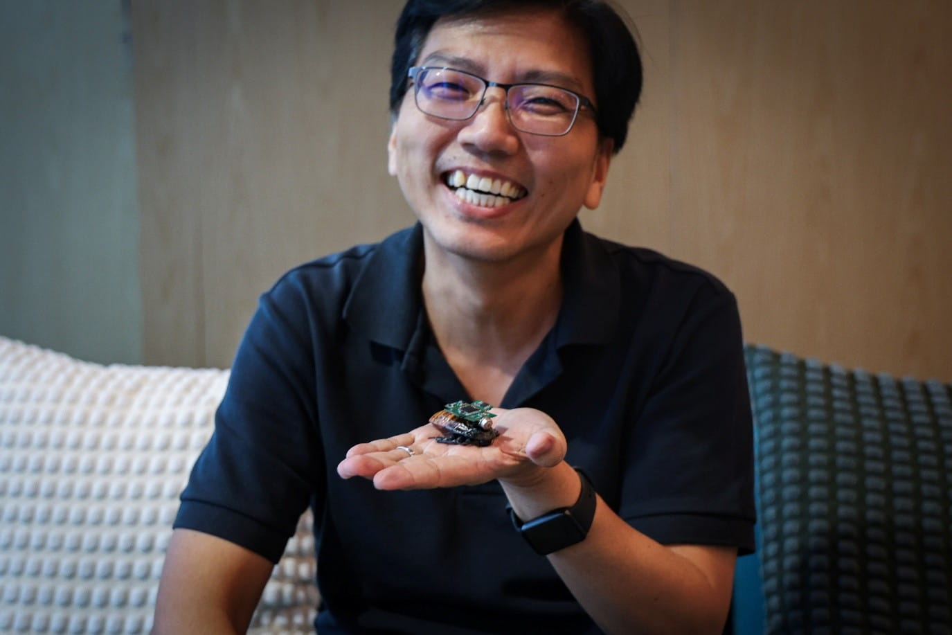 Smiling man seated on a couch presenting a cyborg cockroach on an open palm.