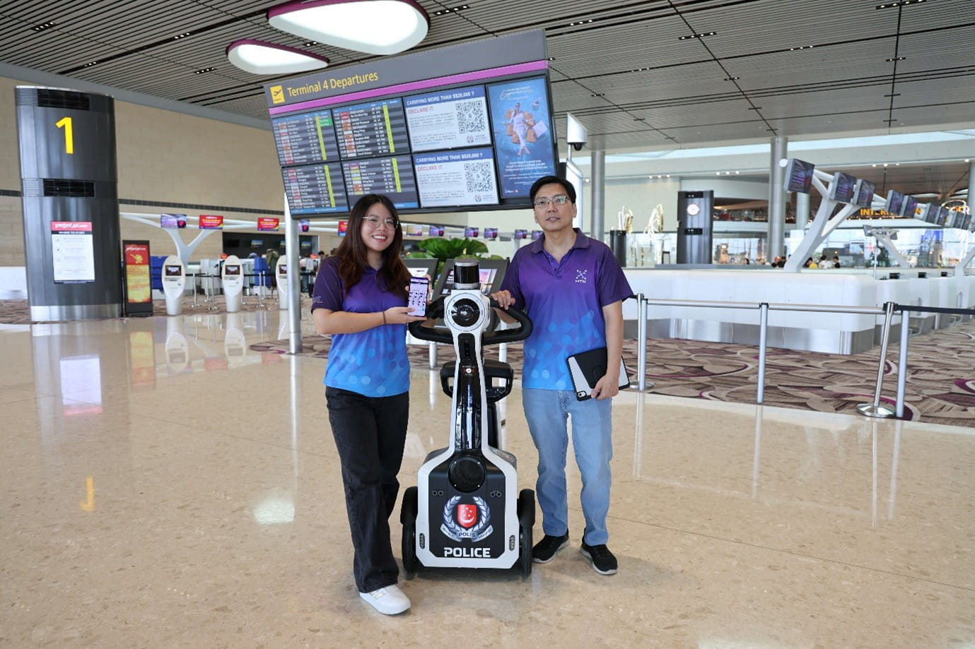 Two people in purple HTX shirts stand with a patrol robot inside an airport terminal.