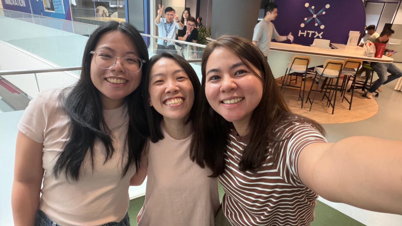 Three women share a joyful selfie in a brightly lit office space.