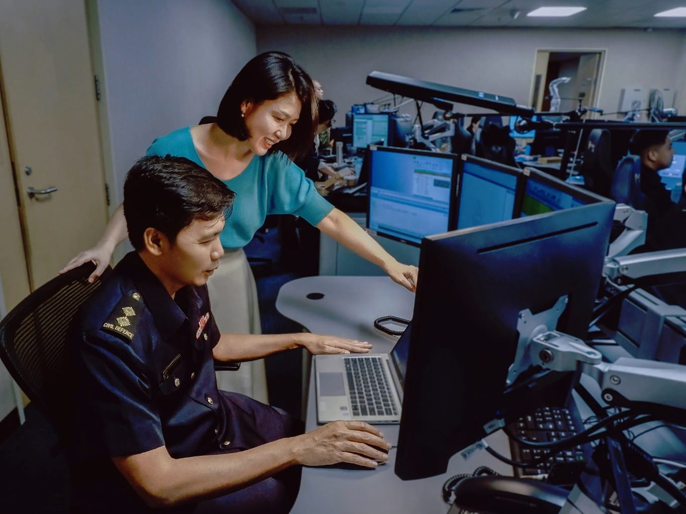 Two people working together in an office setting, with a man seated in a uniform and a woman standing and gesturing at a laptop screen.